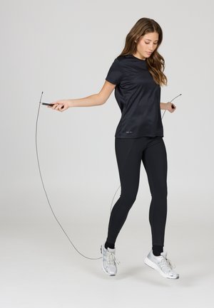 Jonge vrouw in zwarte sportkleding touwtje springend, halverwege een sprong, tegen een effen grijze achtergrond.