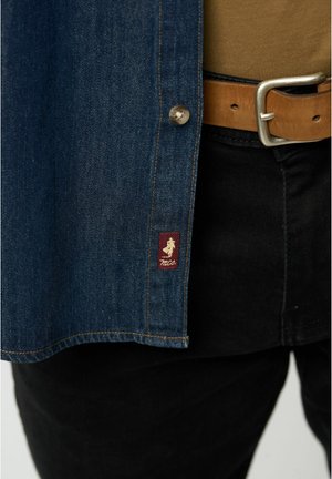 Jeans srajca v temno modri barvi, s kovinskimi gumbom in bordo oznako z logotipom. Nosi se čez rjavo srajco in črne hlače, zavezana z svetlo rjitim pasom.