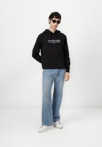 Svart hoodie med framficka och logotyp, kombinerad med ljusblå utsvängda jeans och vita sneakers. Modellen bär också mörka solglasögon.