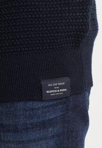 Mörkblå texturerad tröja med ribbad nederkant, med en etikett som säger "ALL DAY WEAR från SCOTCH & SODA, tillverkad med kärlek."