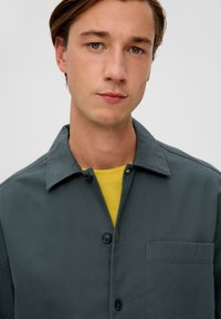 Dunkelgrünes Hemd mit Knopfleiste und Kragen, einer Brusttasche und einem gelben Shirt darunter, mit einer glatten Textur und einfachem Design.