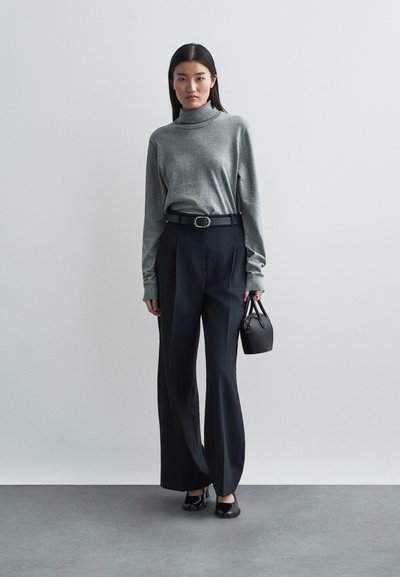 Gray turtleneck sweater, high-waisted black pleated trousers, black belt with round buckle, holding small black handbag, flat black shoes.