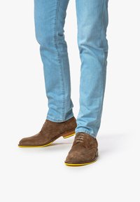 Bruine suede brogue schoenen met gele rubberen zolen, gecombineerd met lichtblauwe slim-fit jeans. Opvallende wingtip-details op de schoenen.