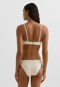 Kremowy komplet bikini o fakturowanym, kropkowanym wzorze. Cienkie, regulowane ramiączka i zapięcie na klips z tyłu. Dół o wysokim stanie.