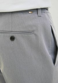 Sive hlače s teksturom koje imaju vidljivi crni gumb, jedan džep s uvučenim detaljima i suptilni hardverski naglasak blizu struka.