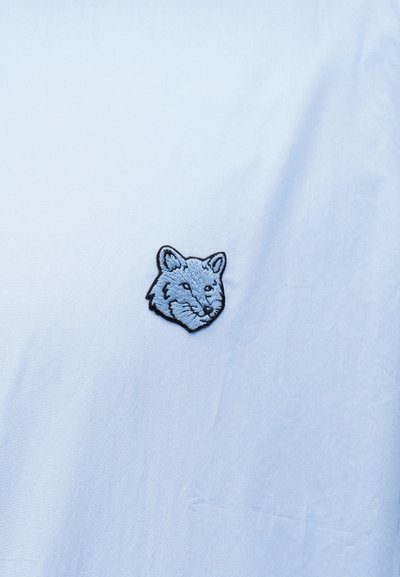 Μπλε κεντημένος λεκές κεφαλής αλεπούς σε ανοιχτό μπλε υφασμάτινο φόντο με διακριτικές ρυτίδες.