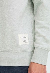 Felpa grigia realizzata in un tessuto morbido, con polsini e orlo a costine, dotata di un'etichetta bianca quadrata con la scritta "11 PROJECT" cucita sul lato.