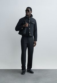 Uomo che indossa giacca nera, pantaloni neri, scarpe nere, occhiali da sole e porta una borsa nera a tracolla, in piedi contro uno sfondo chiaro semplice.