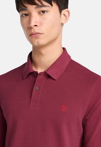 Burgundská polo tričko s texturovanou tkaninou, límečkem, dvěma černými knoflíky a malým červeným logem stromu na levé straně hrudi.