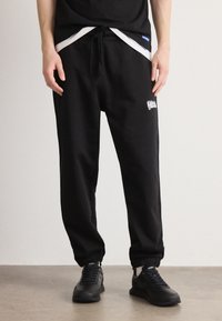 Schwarze Jogginghose mit Kordelzug an der Taille, am Knöchel gesäumt. Verfügt über ein weißes Logo an der Seite und wird mit schwarzen Schnürschuhen kombiniert.