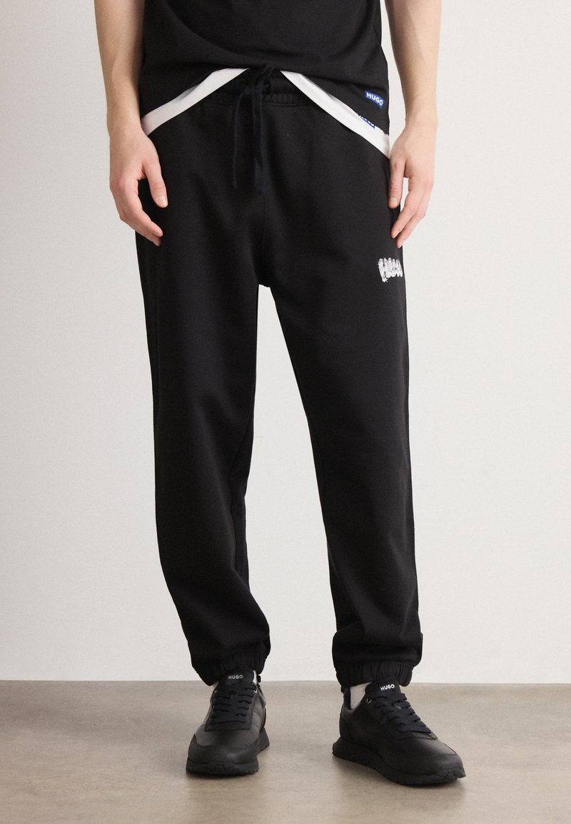 Schwarze Jogginghose mit Kordelzug an der Taille, am Knöchel gesäumt. Verfügt über ein weißes Logo an der Seite und wird mit schwarzen Schnürschuhen kombiniert.