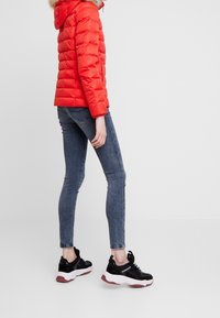 Rød polstret jakke med hætte og pelskant, parret med tætsiddende blå jeans og sorte sneakers med hvide accenter og en tyk sål.