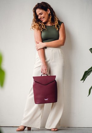 Burgundské kožené batoh s klopou a držadlem. Obsahuje diskrétní logo a strukturovaný tvar, zkombinovaný s béžovými širokými kalhotami.