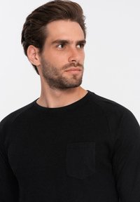 Giovane uomo con capelli corti castani e barba che indossa una maglietta nera a girocollo con una tasca sul petto, che guarda verso destra su uno sfondo bianco semplice.