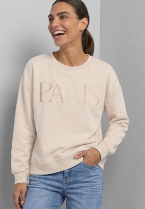 Kvinde smiler og har en lysebeige sweatshirt med ordet "PARIS" broderet, kombineret med blå jeans, stående mod en grå væg.