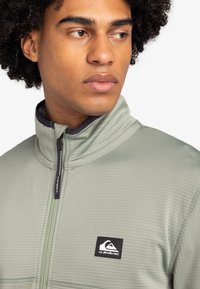 Flísová bunda svetlozeleného odtieňa, s vysokým golierom a textúrovaným povrchom. Náplet s logom Quiksilver na ľavej strane hrude a čierny zips.
