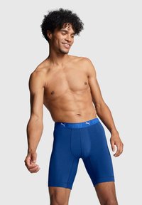 Ablaktalan, göndör hajú férfi kék Puma boxer alsóneműben, egy világos szürke háttér előtt áll.