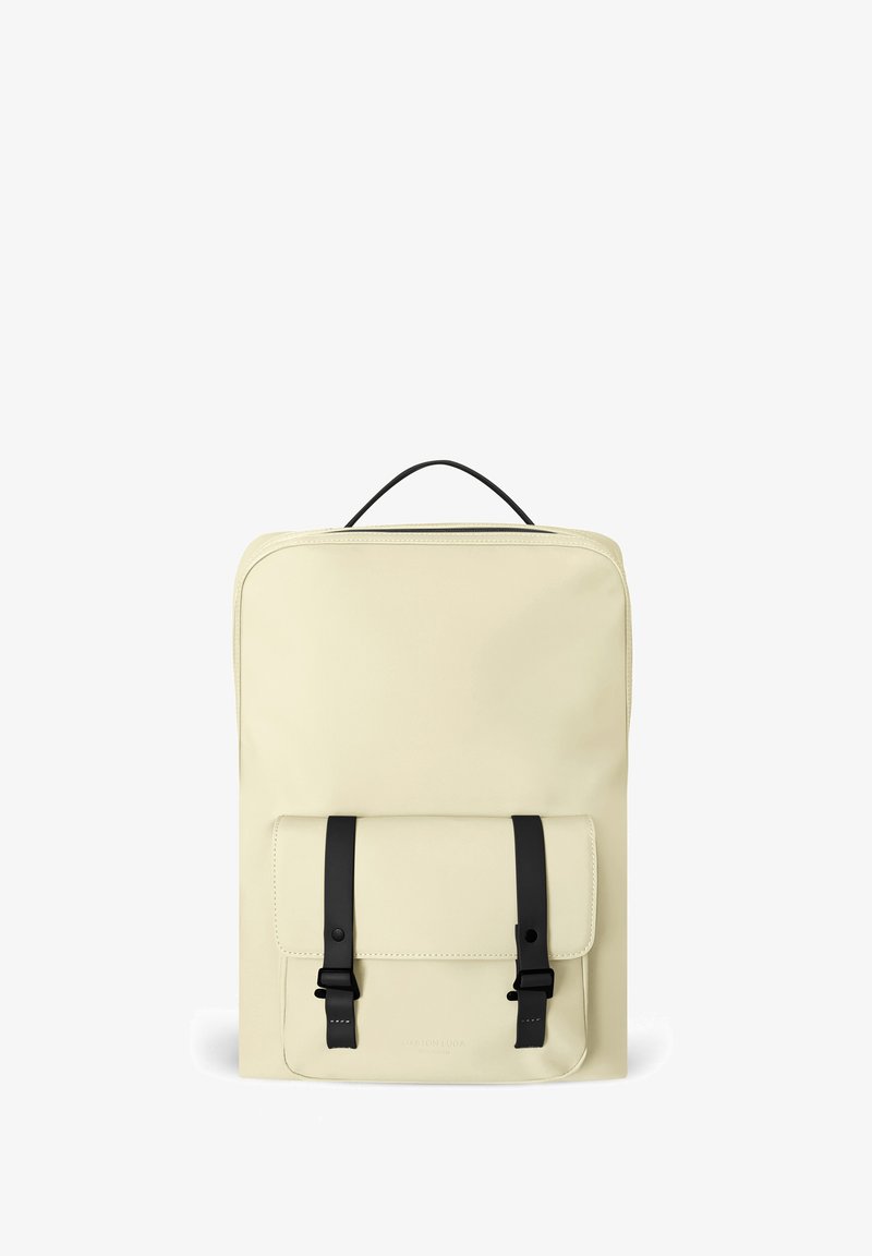 Sac à dos rectangulaire beige avec une poche avant sécurisée par deux sangles noires et une poignée supérieure.