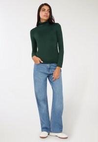 Ciemnozielony golf z długim rękawem w połączeniu z jasnoniebieskimi, wysokimi spodniami w szerokim kroju i białymi sneakersami. Gładki materiał, dopasowany krój.