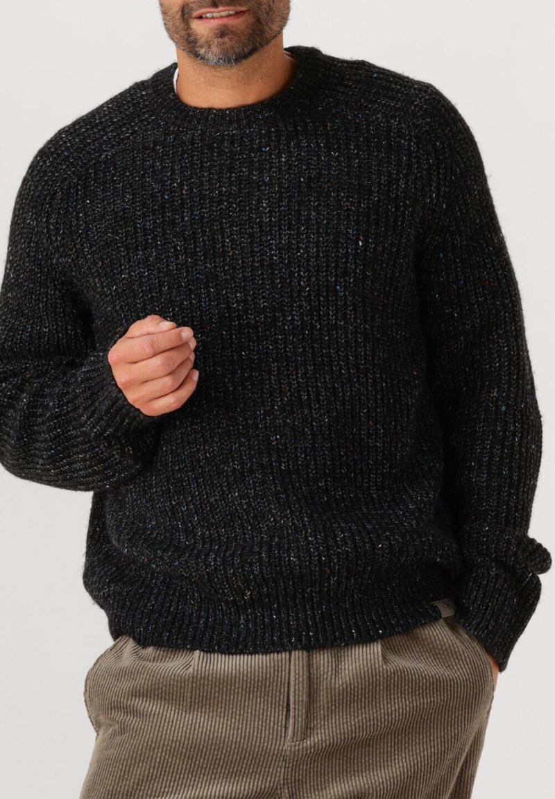 Man in een zwart grof gebreid sweater en beige corduroy broek, met één hand in de zak en de andere omhoog bij de borst.