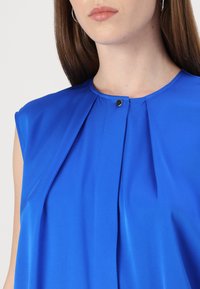 Blusa senza maniche blu con scollatura rotonda, con dettagli plissettati nella parte anteriore e chiusura a bottoni a pressione sul colletto. Tessuto morbido.