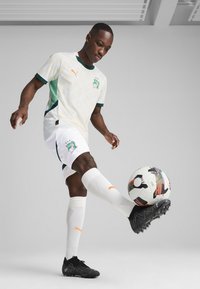 Tenue de football Puma avec un haut crème aux accents verts et un motif géométrique, un short blanc, des crampons noirs et un ballon de football multicolore.