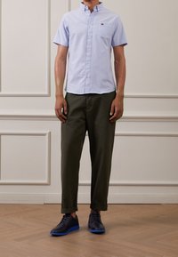 Camicia azzurra a maniche corte con bottoni e taschino sul petto, abbinata a pantaloni verde oliva e scarpe navy con dettagli blu.