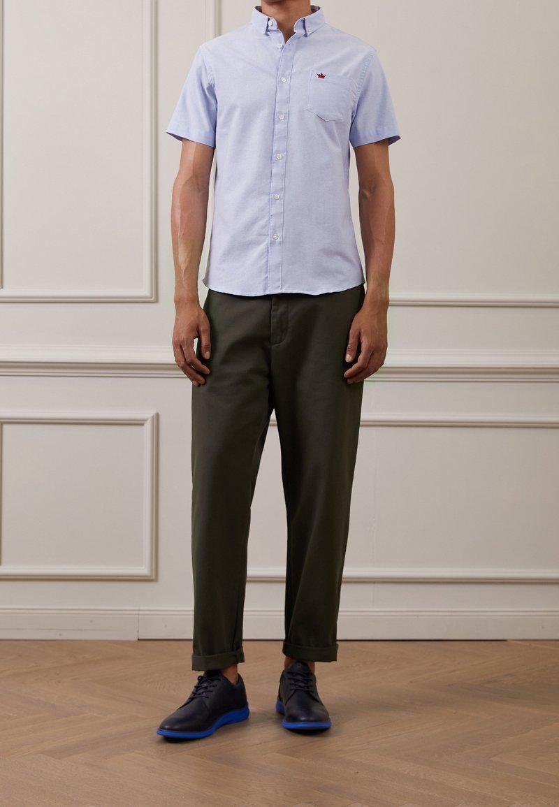 Camicia azzurra a maniche corte con bottoni e taschino sul petto, abbinata a pantaloni verde oliva e scarpe navy con dettagli blu.