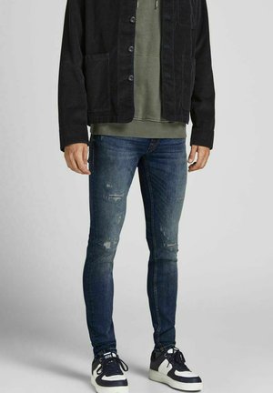 Chaqueta de pana negra, sudadera verde, jeans ajustados azules desgastados y zapatillas negras con acentos blancos. Conjunto casual en capas.