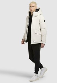 Puffjakke i lys beige med hætte og markante syninger, parret med sorte bukser og hvide sneakers. Har store forlommer.