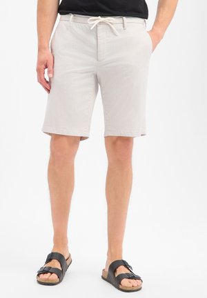 Man wearing light beige drawstring shorts, black T-shirt, and black double-buckle sandals with cork soles, standing with hands in pockets.