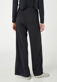 Persona vistiendo pantalones negros holgados y una camiseta de manga larga, de pie descalzo frente a un fondo claro y liso.