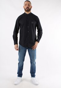 Camicia nera con colletto rialzato e logo sottile, abbinata a jeans azzurri e sneakers bianche su uno sfondo neutro.