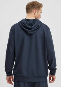 Námornícky modrá mikina s kapucňou vyrobená z mäkkého materiálu, s veľkou kapucňou, dlhými rukávmi a rebrovanými manžetami a lemom. Zadný pohľad ukazuje hladkú textúru.