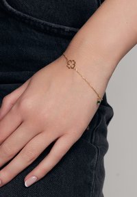 Bransoletka w złotym kolorze z zawieszką w kształcie kwiatu oraz małą zieloną koraliką, na tle ciemniejszej, teksturowanej powierzchni.