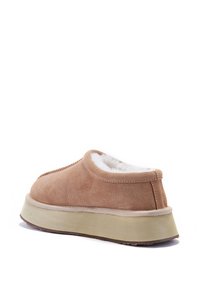 Chaussure en suède beige avec une doublure en fourrure blanche douce, au design arrondi et à semelle rembourrée beige. L'arrière présente un talon légèrement surélevé.