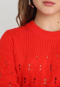Sytě červený pletený svetr s žebrovaným výstřihem a krajkovým vzorem. Charakterizuje ho texturovaná látka a volný, uvolněný střih.