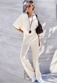 Béžový pletený dvojdielny komplet s širokými nohavicami a voľným topom. Doplnky zahŕňajú okrúhle slnečné okuliare a hnedú kabelku. Súpravu dopĺňajú biele tenisky.