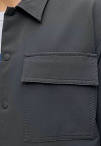 Close-up van het zakje van een donkergrijze jas met klep en zwarte drukknopen op de kraag en het borstgedeelte van een shirt.
