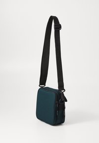 Bolso cruzado cuadrado en color verde azulado con una correa ajustable negra, cierre de cremallera y textura de tela suave. Diseño minimalista sin patrones adicionales.