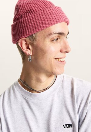 Giovane con berretto rosa lavorato a maglia, orecchini a cerchio argentati, doppia collana d’argento e felpa Vans grigio chiaro, sorride di lato.