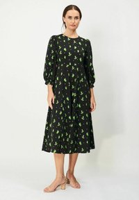 Mustapuku, jossa on vihreä kukkakuosi, lyhyet puffihihat ja liehuva hame. Yhdistettynä beigenvärisiin sandaaleihin. Silkkinen kankaan rakenne.