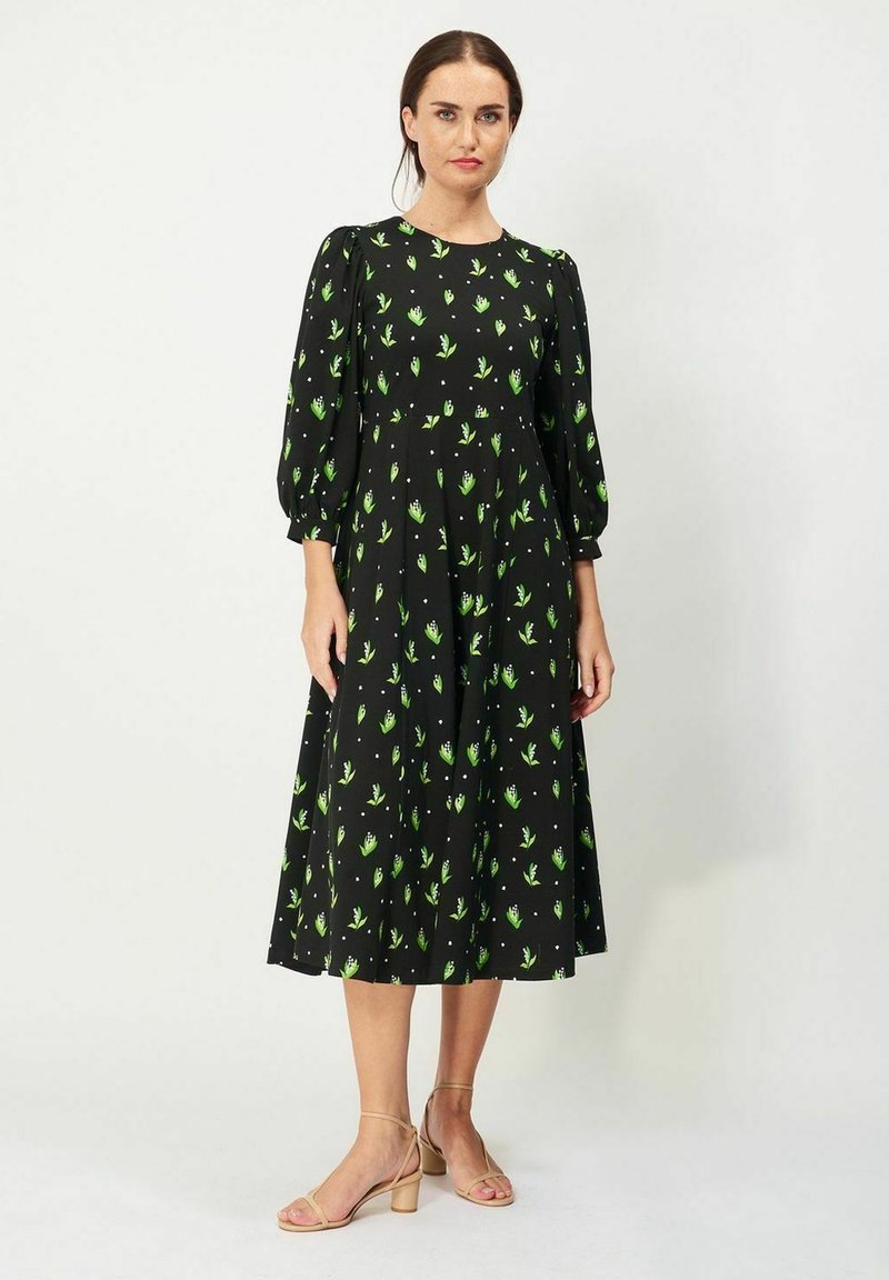 Mustapuku, jossa on vihreä kukkakuosi, lyhyet puffihihat ja liehuva hame. Yhdistettynä beigenvärisiin sandaaleihin. Silkkinen kankaan rakenne.