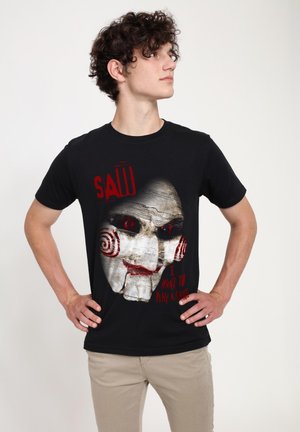 Camiseta de algodón negra con un gráfico de la cara de un títere con ojos rojos y espirales, con la leyenda "SAW QUIERO JUGAR UN JUEGO" en texto rojo.