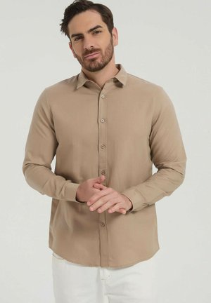 Homme aux cheveux foncés et à la barbe, portant une chemise beige boutonnée et un pantalon blanc, debout, les mains jointes devant un fond clair uni.