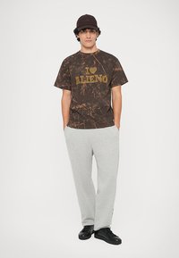 Bruine T-shirt met marmerpatroon en gele tekst "I ❤️ Alieno", gedragen met lichtgrijze joggingbroek en zwarte sneakers. Zwarte pet op het hoofd.