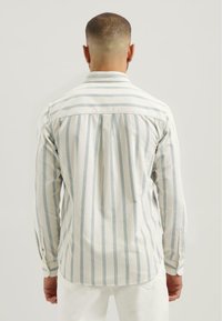 Mężczyzna widziany z tyłu, ubrany w jasny, pionowo prążkowany koszulę z długim rękawem oraz białe spodnie, na jednolitym tle.