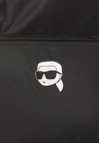 Juodas audinys su lygiu tekstūru pristato išskirtinį baltą ir juodą komiksų veido logotipą, dėvintį akinius nuo saulės, kuris yra šalia užtrauktuko.