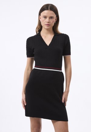 Kvinde iført en sort kortærmet strikket kjole med V-hals og røde, hvide stribede talje- og ærmekanter, stående foran en hvid baggrund.