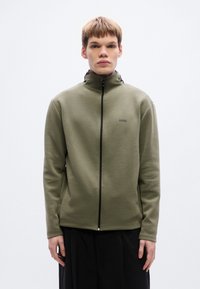 Ελιά πράσινο μπουφάν με φερμουάρ, ψηλός γιακάς, μακριά μανίκια και μινιμαλιστικό σχέδιο· διαθέτει μαύρο λογότυπο στο αριστερό στήθος και τσέπες με φερμουάρ.